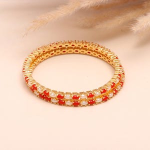 May include: Two gold bangles with alternating red and white stones. The bangles are circular and have a textured gold band. The red stones are a vibrant colour, and the white stones appear to be pearls. The background is a light beige colour.