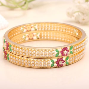 Könnte beinhalten: Zwei goldene Armreifen mit weißen Kunstperlen und floralen Akzenten. Die Armreifen weisen ein sich wiederholendes Muster aus kleinen Perlen auf und sind mit roten und grünen floralen Designs verziert. Wunderschöner Schmuck.