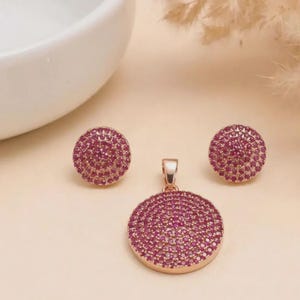 May include: A jewellery set consisting of a circular pendant and two matching circular stud earrings. Each piece is crafted from rose gold-toned metal and features a dense, pavé-style setting of small, vibrant pink stones covering the entire surface.