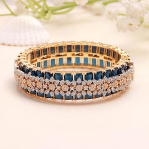 May include: Gold-coloured bangle bracelet featuring teal rectangular stones and clear, round-cut stones. The bracelet has a floral pattern in the centre. The background is a soft, neutral colour.