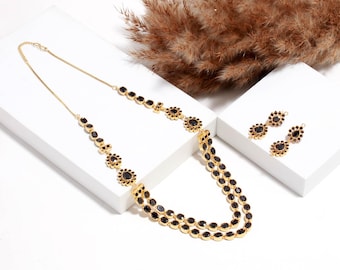 Könnte beinhalten: Ein goldfarbenes Halsketten- und Ohrring-Set. Die Halskette hat zwei Stränge aus schwarzen, oval geformten Steinen und floralen Akzenten. Die Ohrringe haben ein ähnliches Design mit schwarzen Steinen und goldfarbenen Fassungen. Das Schmuckstück wird auf einer weißen Oberfläche präsentiert.