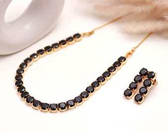 Könnte beinhalten: Ein goldfarbenes Halsketten- und Ohrring-Set. Die Halskette und die Ohrringe weisen eine Reihe schwarzer Edelsteine auf. Der Schmuck wird auf einer weißen Oberfläche präsentiert, mit einer dekorativen weißen Vase und Trockenblumen im Hintergrund.