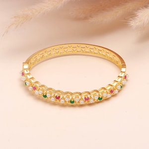 May include: Gold-coloured bangle bracelet with a decorative design. The bracelet features a row of small white pearls, red and green gemstones. The top of the bracelet has a geometric pattern.