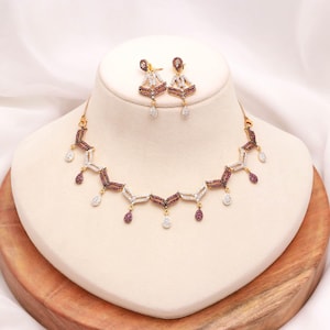 Könnte beinhalten: Eine goldfarbene Halskette und ein passendes Paar Ohrhänger mit einem sich wiederholenden Chevron-Muster. Der Schmuck ist mit abwechselnd dunkelroten und klaren weißen Steinen besetzt, wobei kleine tropfenförmige Anhänger an den Gliedern hängen.