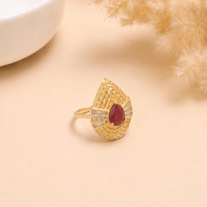 Peut inclure: Une bague en or avec une pierre précieuse rouge en forme de poire au centre, entourée d'une monture en or texturée en éventail. Le design comprend de petites pierres claires taille baguette disposées en éventail autour de la pierre rouge.