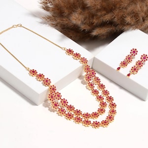 May include: A gold-toned necklace and matching earrings set. The necklace features a layered design with red flower-shaped accents. The earrings have a similar floral design. The jewelry set is displayed on a white surface.