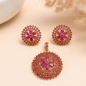 May include: A jewelry set consisting of two round stud earrings and a matching pendant. Each piece features a central flower motif made of six pear-shaped red stones, surrounded by multiple concentric circles of tiny red stones set in gold.