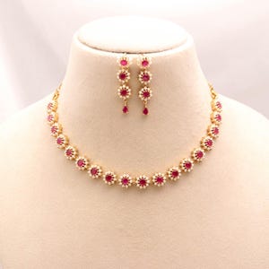 May include: A gold-toned necklace and matching earring set featuring a series of flower-shaped links. Each link contains a central round red stone surrounded by small white stones. The earrings mirror the floral design with a single drop element.