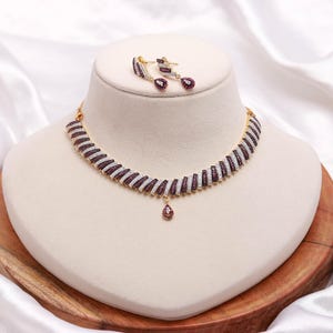 Puede incluir: Un conjunto de collar y pendientes en tono dorado con un patrón repetitivo de filas diagonales. Cada fila alterna entre piedras blancas transparentes y piedras de color rojo intenso. El collar incluye un colgante central en forma de gota.