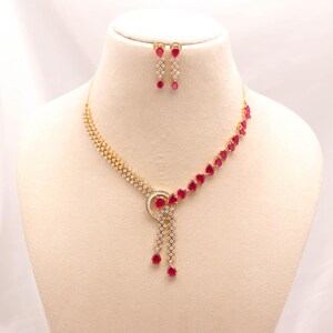 Puede incluir: Un conjunto de collar y pendientes colgantes en tono dorado con piedras rojas en forma de lágrima y cristales blancos transparentes. El collar presenta un motivo circular central con dos hilos colgantes, a juego con los pendientes.