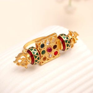 Può includere: Un anello d'oro con un elemento centrale quadrato e due estremità decorative. Il quadrato presenta pietre preziose colorate. Le estremità sono rosse e verdi con dettagli dorati. L'anello è su una superficie bianca.