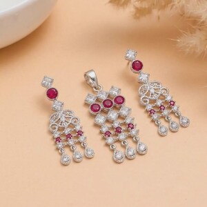 Puede incluir: Un conjunto de joyería compuesto por un colgante y dos pendientes a juego, fabricado en metal plateado. Cada pieza presenta piedras redondas de color rojo intenso y piedras transparentes cuadradas en un diseño colgante de filigrana.