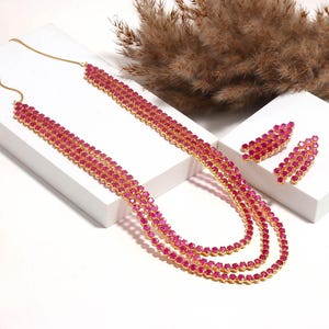 Puede incluir: Un conjunto de collar de tres hilos y pendientes a juego. La joyería presenta pequeñas piedras redondas de color rubí engastadas en un metal dorado. El collar está drapeado sobre una superficie blanca, y los pendientes se exhiben en un bloque blanco.
