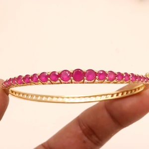 May include: A gold-toned hinged bangle bracelet featuring a row of round, vibrant fuchsia-pink gemstones set along the top. The metal band has a polished finish and a secure clasp mechanism, showcasing a series of uniform stones in a line.