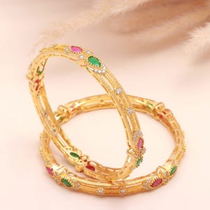 May include: Two gold bangles with detailed patterns. Each bangle has alternating red, green, and clear gemstones, plus small, sparkling details. The bangles are presented on a soft, light-coloured surface.
