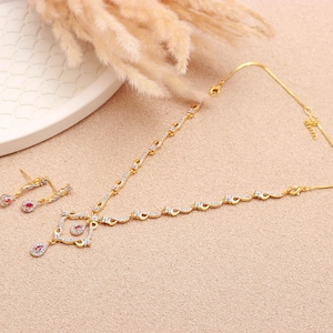 Puede incluir: Un conjunto de joyas de tono dorado con un collar y pendientes a juego. El collar tiene un colgante cuadrado central con una lágrima colgante, y los pendientes son diseños pequeños y colgantes. Ambas piezas están adornadas con piedras preciosas claras y rojas.