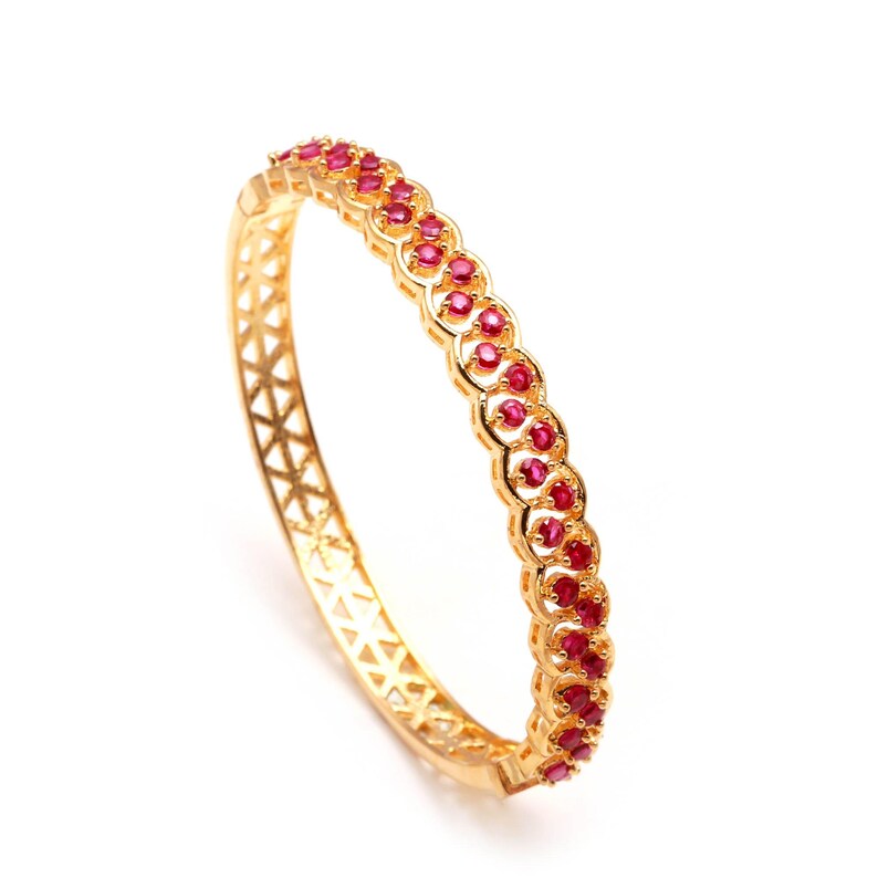 K&ouml;nnte beinhalten: Ein goldfarbenes Armreif-Armband mit einem sich wiederholenden Muster aus kleinen, runden, roten Edelsteinen. Das Armband hat ein dekoratives, durchbrochenes Design an den Seiten.