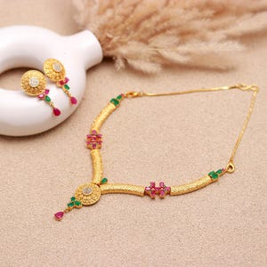 May include: A gold-toned jewelry set featuring a necklace and matching earrings. The necklace has a textured band with red and green gemstone accents. The earrings are round with a central design and dangling gemstones. The set is displayed on a neutral surface.