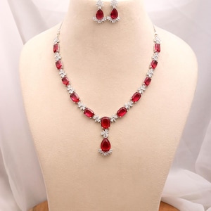 May include: A jewelry set consisting of a necklace and matching earrings. The necklace features rectangular red stones alternating with small clear stones, ending in a teardrop-shaped red stone pendant. The earrings are matching red teardrop stones.