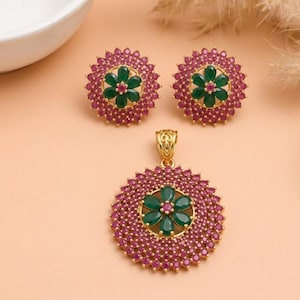 Könnte beinhalten: Ein Schmuckset bestehend aus einem Anhänger und zwei passenden Ohrringen. Jedes Stück verfügt über eine goldfarbene, kreisförmige Basis, die mit zahlreichen kleinen rosa Steinen besetzt ist, welche ein zentrales Blumenmotiv umgeben.