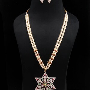 May include: A pearl necklace and matching earrings set. The necklace has a double strand of pearls with red and gold accents, and a large star pendant with clear and coloured gemstones. The earrings are small, diamond-shaped, and adorned with gemstones.