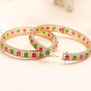 Peut inclure: Deux bracelets dorés ornés de pierres carrées vertes et roses alternées. Les bracelets ont un fermoir à charnière et sont sertis de petites pierres claires sur les bords. Les bijoux sont présentés sur un fond clair.
