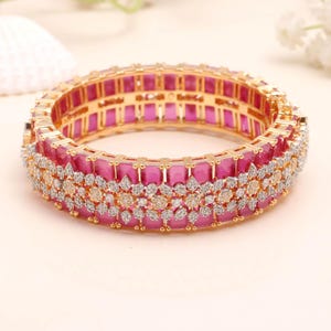 Puede incluir: Una pulsera de brazalete dorada con un centro de piedras preciosas de color rosa vibrante y un diseño ornamentado. La pulsera presenta una fila de piedras rosas rectangulares, acentuadas por una capa de piedras más pequeñas, claras y doradas.