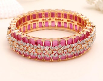 May include: A gold-coloured bangle bracelet with a vibrant pink gemstone centre and an ornate design. The bracelet features a row of rectangular pink stones, accented by a layer of smaller, clear and gold-coloured stones.