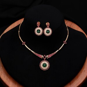 May include: A gold necklace and earring set. The necklace has a pendant with a green centre, white beads, and red accents. The earrings match the pendant design. The set is displayed against a black backdrop.