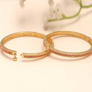 Op de afbeelding: Twee goudkleurige armbanden met een rij kleine rode edelstenen. De armbanden zijn open, waardoor het sluitmechanisme zichtbaar is. Zachte bloemaccenten op de achtergrond voegen elegantie toe.