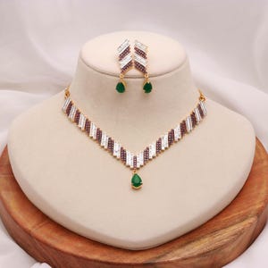Könnte beinhalten: Ein goldfarbenes Halsketten- und Ohrringset mit V-förmigem Design. Der Schmuck ist mit Reihen aus klaren Baguette-Steinen und dunkelvioletten Steinen verziert und wird durch einen tropfenförmigen grünen Steinanhänger ergänzt.
