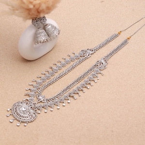 May include: A silver-toned necklace with an ornate pendant and matching earrings. The necklace features a detailed design with a central pendant and a delicate chain. The earrings are bell-shaped with intricate detailing. The jewelry is displayed on a neutral-toned surface.