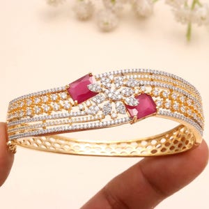 Puede incluir: Un brazalete rígido articulado en tono dorado con dos piedras rojas de corte rectangular y un motivo floral central incrustado con numerosas piedras pequeñas, claras y brillantes. La banda tiene un patrón de rejilla y acabado dorado.