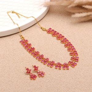 Puede incluir: Un conjunto de collar y pendientes dorados. La joyería presenta piedras preciosas rosas rectangulares engastadas en un marco dorado. El conjunto se exhibe sobre una superficie neutra.