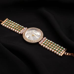 Puede incluir: Reloj de pulsera dorado con esfera redonda, rodeada de pequeños cristales transparentes. La correa presenta filas de perlas blancas y piedras verdes. El reloj está sobre un fondo negro.