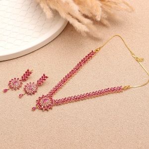 May include: A gold-toned jewelry set featuring a necklace and matching earrings. The necklace and earrings are adorned with vibrant red gemstones arranged in a floral pattern. The set is displayed on a neutral-toned surface.