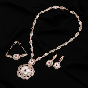 Puede incluir: Un conjunto de joyería que incluye un collar, una pulsera y un par de pendientes colgantes. Cada pieza presenta metal en tono oro rosa, pétalos de flor de nácar blanco y pequeñas piedras rosas engastadas en un patrón de filigrana.