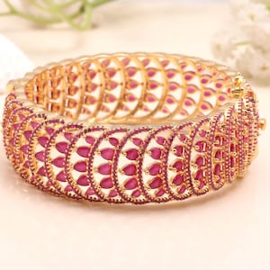 May include: A gold-plated bangle bracelet featuring a repeating pattern of ruby-red gemstones. The gemstones are teardrop-shaped and set in an openwork design. This bracelet is a striking piece of jewellery.
