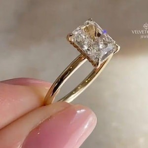 Könnte beinhalten: Ein goldener Verlobungsring mit einem rechteckig geschliffenen Diamanten. Der Diamant hat eine strukturierte Oberfläche und ist auf einem zarten Goldband gefasst. Der Hintergrund ist unscharf. Das Logo "VELVETGLOW JEWELS" ist sichtbar.