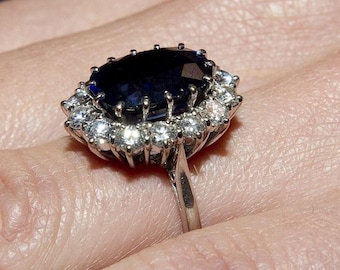 Op de afbeelding: Een zilveren ring met een grote, ovale, donkerblauwe edelsteen omringd door kleinere, heldere edelstenen. De ring heeft een eenvoudige zilveren band. De edelstenen zijn gezet in een halo-ontwerp, wat een sprankelend effect creëert.