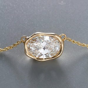 Könnte beinhalten: Eine goldene Halskette mit einem oval geschliffenen Diamantanhänger. Der Diamant ist in einer Lünettenfassung gefasst und hängt an einer feinen Goldkette. Der Anhänger ist der Blickfang und unterstreicht das Funkeln des Diamanten vor neutralem Hintergrund.