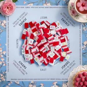 May include: A mahjong set featuring red and white tiles with floral and graphic designs, arranged on a light blue play mat labeled with cardinal directions and game rules. The mat includes text regarding jokers and tile sequences.