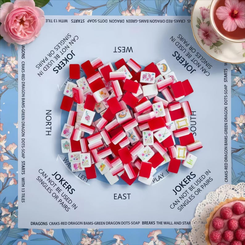 May include: A mahjong set featuring red and white tiles with floral and graphic designs, arranged on a light blue play mat labeled with cardinal directions and game rules. The mat includes text regarding jokers and tile sequences.