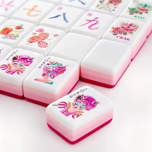May include: A set of rectangular mahjong tiles with white tops and layered pink sides. The tiles feature colorful illustrations of characters with pink hair, floral motifs, and Chinese numerals. One tile in the foreground is labeled Joker.