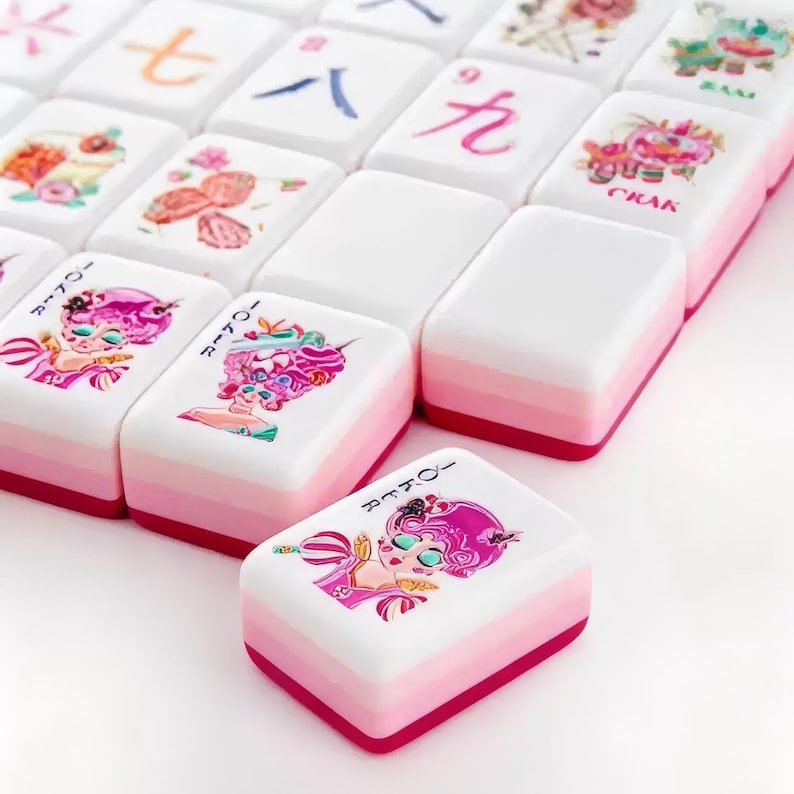 May include: A set of rectangular mahjong tiles with white tops and layered pink sides. The tiles feature colorful illustrations of characters with pink hair, floral motifs, and Chinese numerals. One tile in the foreground is labeled Joker.