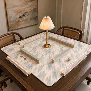 May include: A square mahjong mat with a light blue and cream abstract pattern, featuring four long tile racks arranged in a square formation. A brass-toned table lamp with a pleated white shade sits in the center of the mat on a dark wood table.