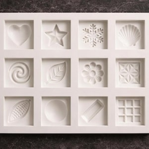Può includere: Uno stampo in silicone bianco con dodici scomparti quadrati, ciascuno con un diverso disegno in rilievo. I disegni includono un cuore, una stella, un fiocco di neve, una conchiglia, un vortice, una foglia, un fiore e forme geometriche.