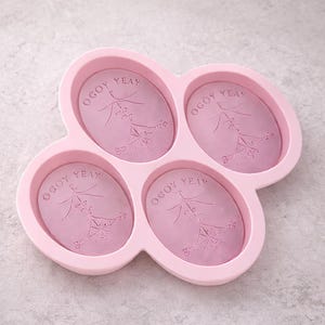Puede incluir: Un molde de jab&oacute;n de silicona rosa con cuatro cavidades ovaladas. Cada cavidad tiene el texto "OGOY YEAY" y un dise&ntilde;o floral decorativo. El molde est&aacute; dise&ntilde;ado para hacer jabones artesanales.