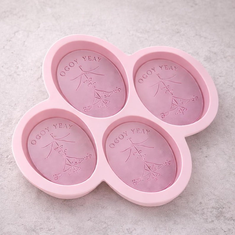 Puede incluir: Un molde de jab&oacute;n de silicona rosa con cuatro cavidades ovaladas. Cada cavidad tiene el texto "OGOY YEAY" y un dise&ntilde;o floral decorativo. El molde est&aacute; dise&ntilde;ado para hacer jabones artesanales.