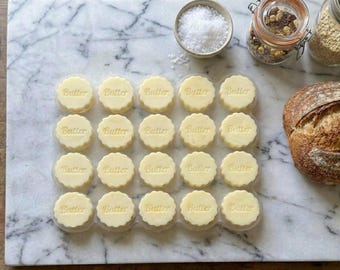 May include: A top-down shot of a marble surface with individual portions of butter arranged in a grid. Each butter portion is round with a scalloped edge and the word "Butter" embossed on top. A loaf of bread, salt, and nuts are also visible.
